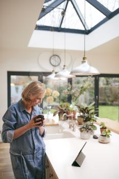 Mature Woman At Home In Kitchen Drinking Coffee And Looking At Digital Tablet