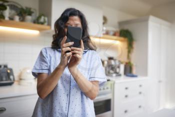 Mature Hispanic Woman In Pyjamas At Home In Kitchen Texting And Using Social Media On Mobile Phone