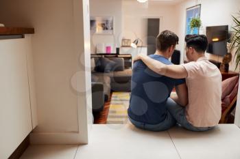 Rear View Of Loving Same Sex Male Couple Hugging As They Relax At Home Together
