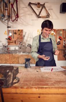 Excited Male Business Owner In Workshop Having Taken Call On Mobile Phone