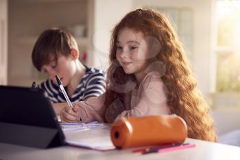 Two Children Sitting At Kitchen Counter Doing Homework Using Digital Tablet