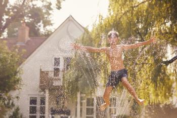 Boy Wearing Swimming Costume Having Fun In Summer Garden Playing In Water From Garden Sprinkler