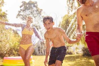 Family Running Through Water From Garden Sprinkler Having Fun Wearing Swimming Costumes