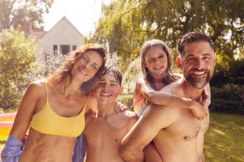 Portrait Of Family Wearing Swimming Costumes Having Fun Playing In Water From Garden Sprinkler