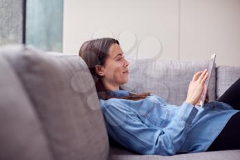 Smiling Young Woman At Home Lying On Sofa Looking At Digital Tablet