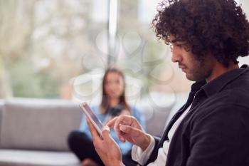 Relaxed Young Couple At Home Sitting On Sofa Using Digital Tablet And Mobile Phone