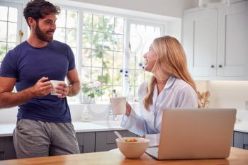 Couple Wearing Pyjamas In Kitchen With Laptop Eating Breakfast Together