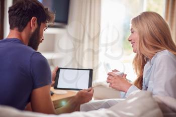 Couple Wearing Pyjamas In Lounge At Home Chatting And Using Digital Tablet