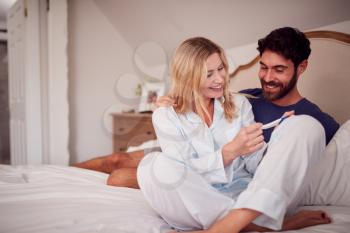 Excited Couple Wearing Pyjamas In Bedroom Celebrating Positive Pregnancy Test