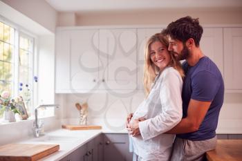Couple With Man Hugging Pregnant Woman Wearing Pyjamas Standing In Kitchen Holding Bump