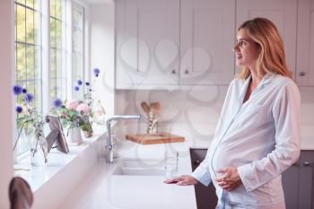 Pregnant Woman Wearing Pyjamas Standing In Kitchen Holding Bump