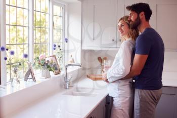 Couple With Man Hugging Pregnant Woman Wearing Pyjamas Standing In Kitchen Holding Bump
