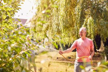 Retired Couple At Work Watering Plants With Hose And Tidying Garden With Rake At Home