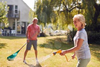 Retired Couple At Work Watering Plants With Hose And Tidying Garden With Rake At Home