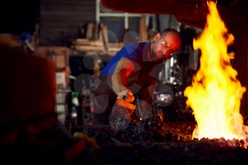 Male Blacksmith Using Wire Brush On Metalwork Heating In Forge