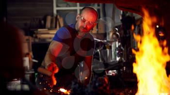 Male Blacksmith Using Wire Brush On Metalwork Heating In Forge