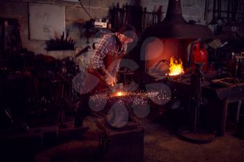 Male Blacksmith Hammering Metalwork On Anvil With Blazing Forge In Background