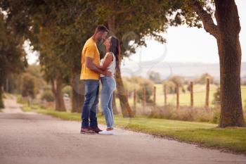 Side View Of Loving Couple Hugging As They Walk Along Track Through Countryside