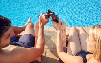 Over The Shoulder View Of Loving Couple On Summer Vacation Drinking Beer In Chairs By Swimming Pool