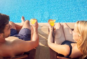 Over The Shoulder View Of Couple On Summer Vacation With Cold Drinks In Chairs By Swimming Pool