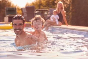 Portrait Of Father And Son Playing In Outdoor Pool On Vacation As Mother And Baby Watch From Side