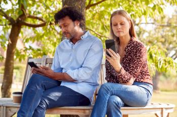 Mature Couple Meeting In Outdoor Park Sitting On Bench Looking At Mobile Phones