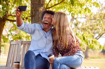 Kissing Mature Couple Posing For Selfie On Mobile Phone Sitting On Seat In Park