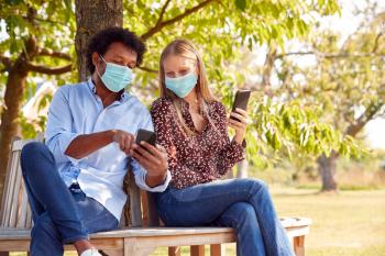 Couple Wearing Masks Meeting In Outdoor Park During Health Pandemic Looking At Mobile Phones