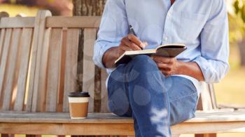 Close Up Of Man With Takeaway Coffee Sitting On Park Bench Under Tree Writing In Notebook