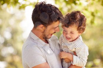 Loving Father With Son Having Fun In Park Holding Him In The Air Against Leafy Background