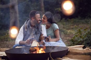 Romantic Couple Camping Sitting By Bonfire In Fire Bowl With Hot Drinks