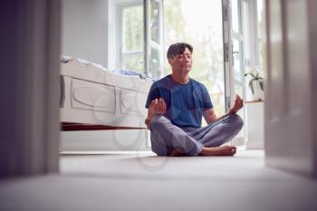 Mature Asian Man In Pyjamas Sitting On Bedroom Floor Meditating In Yoga Pose