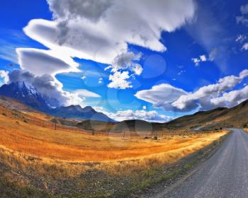 Gray dirt road in the Chile National Park Torres del Paine. Incredible shaped cloud formed by glaciers glisten in the sun. On the horizon are seen mountains with snow-capped peaks. Picture taken wit
