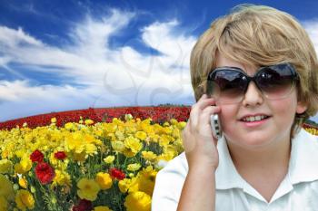 Beautiful blond boy with sunglasses talking on his mobile phone. Behind him, the field of blooming yellow and red buttercups