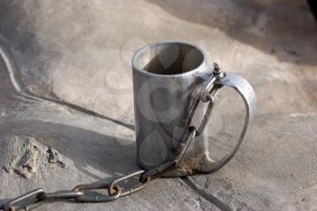  A metal mug for water with a chain near to a source. 
