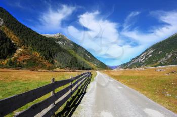 Wide fenced road in an alpine valley. Sunny autumn day. The mountain slopes are covered with dense pine forest