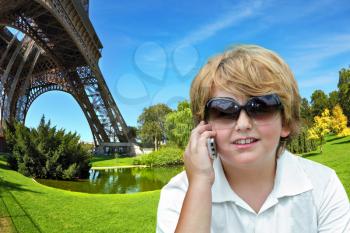 Beautiful blond boy having fun chatting with friends on a cell phone. Background - one of the pillars of the Eiffel Tower