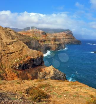 Eastern tip of the island of Madeira. Rocks steeply in the blue waters of the Atlantic Ocean. Over a cliff on the ocean breeze are the windmills