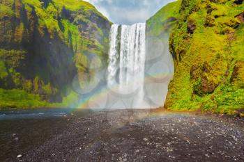 Picturesque huge rainbow appears in the water mist. The most popular waterfall in Iceland - Skogafoss. Water rushes down with a crash, forming a cloud of mist