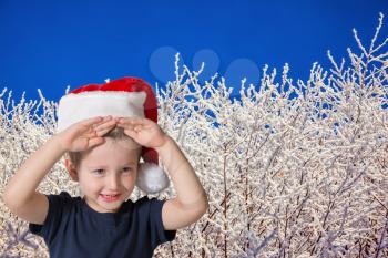 Three year old boy in red cap of Santa Claus. Cute kid having fun smiling on the background of snow-covered forest