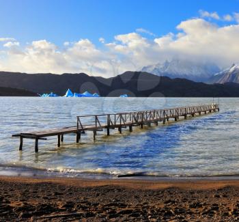 National Park Torres del Paine, Chile. Pier on Lake Grey. In the distance swims picturesque blue iceberg. In the mountains around the lake are piled clouds. Summer sun illuminates the lake.