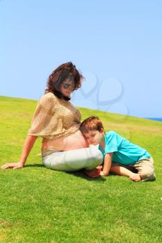 Beautiful young pregnant woman rests with lovely little son on the grass near the sea