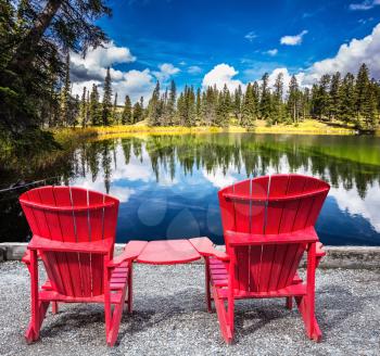  Jasper National Park in the Rocky Mountains. Two red plastic chairs on the lake. On shores of the lake grow coniferous forests