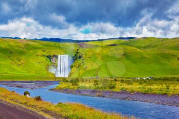  Iceland, waterfall Skogafoll. Huge picturesque waterfall and creek running along the road