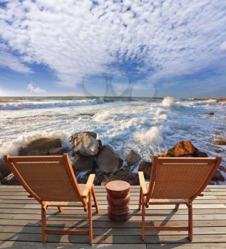Charming lookout for two. Two convenient comfortable guest sun loungers and a bedside table are worth on a wooden platform. It is well-admired stormy sea surf