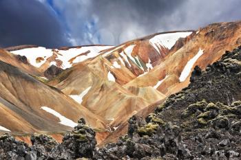 The orange and beige rhyolite mountains with snow in the hollows. National Park Landmannalaugar in Iceland. Pieces of gray and black lava, sometimes covered with green moss.