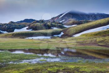  Snow lies in the hollows of colorful rhyolite mountains. Green Valley is flooded with melt water. Early summer morning in the National Park Landmannalaugar, Iceland