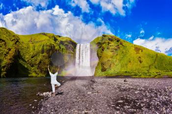  Elderly woman performs asana tree of hatha yoga before falling water. Grand waterfall with rainbow in the water mist Skogafoss