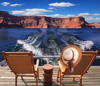 Waves from the boat cut through the Lake Powell. At the stern of the vessel are two deck chairs. On the back of one hanging elegant ladies straw hat.