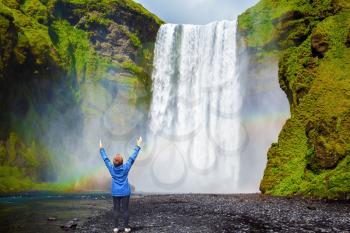 Interesting waterfall in Iceland - Skogafoss. Picturesque huge rainbow appears in the water mist. Middle-aged woman - tourist shocked beauty waterfall